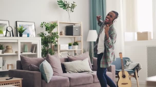 Estudante afro-americano bonito está dançando em casa se divertindo ouvindo música com fones de ouvido. Apartamento luz bonita está no fundo . — Vídeo de Stock