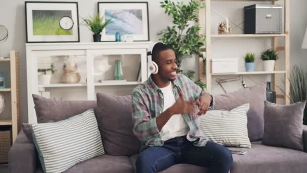 Feliz chico afroamericano está bailando y cantando escuchando música a través de auriculares sentados en el sofá en casa relajándose disfrutando del tiempo libre. Millennials y concepto de felicidad . — Vídeo de stock