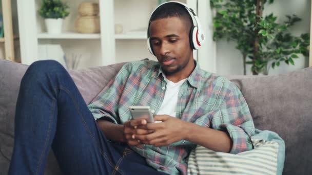 Jovem afro-americano está usando telefone inteligente sentado no sofá no apartamento e ouvindo música através de fones de ouvido. Dispositivos e conceito de estilo de vida . — Vídeo de Stock