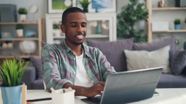 Χαρούμενα αφρικανική αμερικανική guy μακρινό εργαζόμενος ευρίσκεται στην εργασία στο σπίτι, χρησιμοποιώντας φορητό υπολογιστή που κάθεται στο γραφείο στο στούντιο κοιτάζοντας οθόνη δακτυλογράφηση και κάνοντας σημειώσεις. Επιχειρήσεων και νέων concept. — Αρχείο Βίντεο