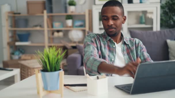 Afrikanisch-amerikanischer Kerl arbeitet zu Hause mit Laptop-Tippen, macht Notizen auf Papier und trinkt am Schreibtisch am Arbeitsplatz in heller Wohnung. — Stockvideo