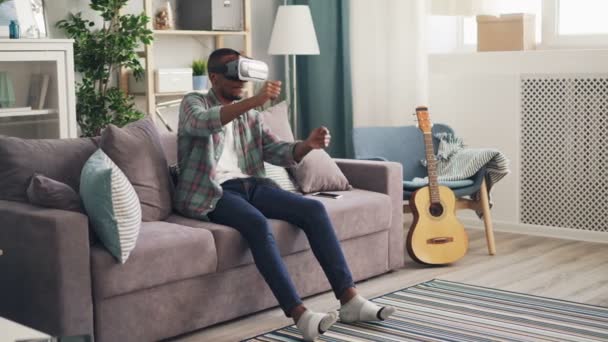 El chico afroamericano moderno está jugando en gafas de realidad virtual conduciendo carreras de coches sentados en un sofá solo y disfrutando del entretenimiento contemporáneo . — Vídeos de Stock