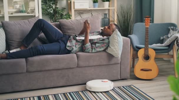 Безтурботний студент прибирає свою кімнату за допомогою роботизованого пилососа і лежить на дивані, розслабляючись і використовуючи смартфон в сучасній квартирі. Молодіжна та побутова концепція . — стокове відео