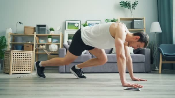 Przystojny mężczyzna ćwiczeń w domu robi ćwiczenia cardio w deski za pomocą smartfona. — Wideo stockowe