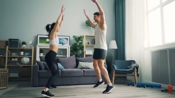 Gente alegre pareja en ropa deportiva haciendo deportes saltando levantando brazos en casa — Vídeo de stock