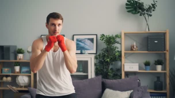 Jovem boxeador em sportswear treinamento em casa boxe sozinho com pulsos envoltos — Vídeo de Stock