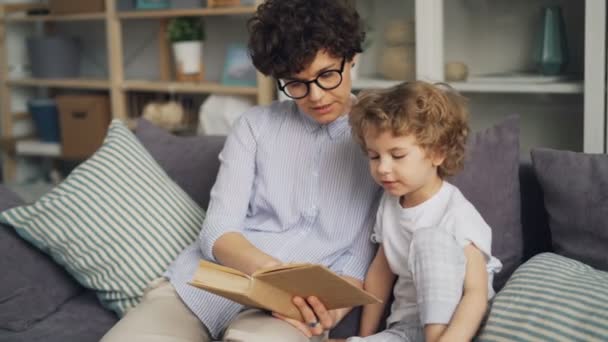 Słodkie małe dziecko czytając książkę z kochającą matką i uśmiecha się na kanapie w mieszkaniu — Wideo stockowe