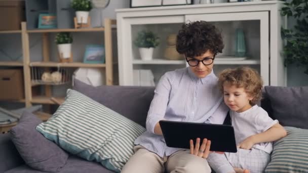 Очаровательный мальчик смотрит мультики на планшете со своей заботливой матерью, держащей гаджет — стоковое видео