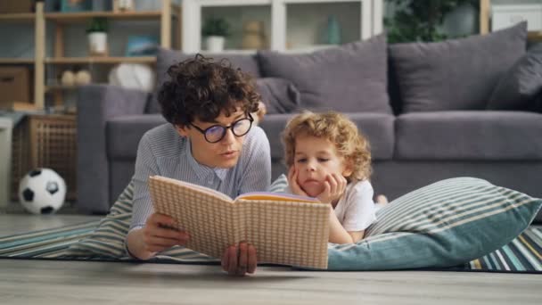 Giovane donna madre lettura libro a bambino sdraiato sul pavimento su una coperta insieme — Video Stock