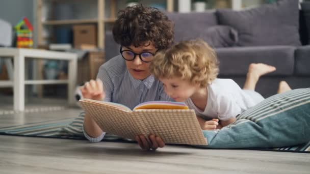 Щаслива родина молода жінка і маленький хлопчик читає книгу, що лежить на ковдрі вдома — стокове відео