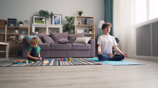 Criança brincando com blocos no chão enquanto sua mãe meditando na posição de lótus — Vídeo de Stock