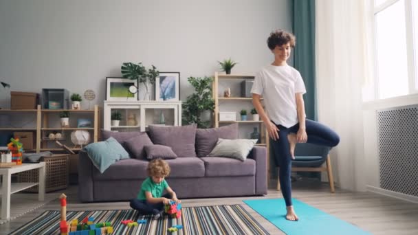 小男孩玩玩具在地板上, 当他的运动的母亲做瑜伽垫 — 图库视频影像