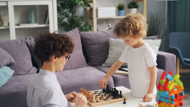 Niño pequeño y su madre jugando al ajedrez en casa y hablando de juego — Vídeo de stock