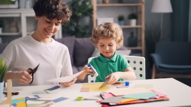 Madre e hijo haciendo collages cortando papel y pegándose con pegamento en la mesa — Vídeos de Stock