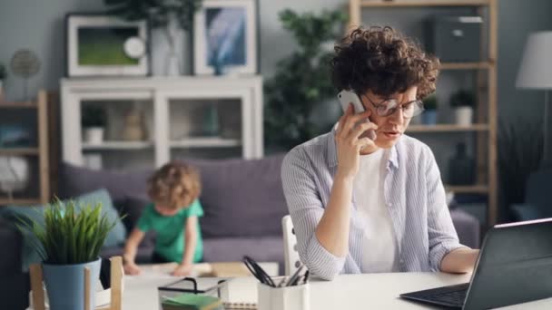 Vztek na volné noze mluví o telefonu pomocí přenosného počítače, zatímco její syn hraje v pokoji — Stock video