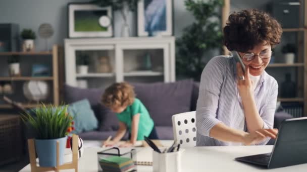 Chica hablando en el teléfono móvil trabajando con el ordenador portátil, mientras que su hijo jugando en casa — Vídeos de Stock