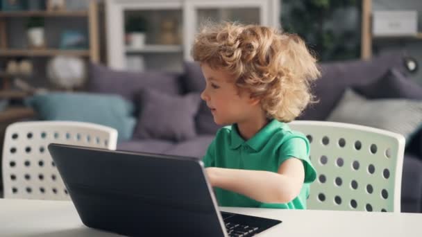 Lindo niño jugando con el ordenador portátil moderno pulsando botones y sonriendo en casa — Vídeos de Stock