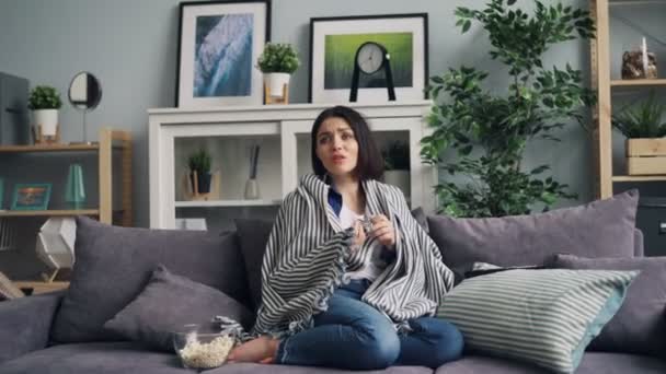 Молодая женщина смотрит страшный триллер по телевизору, сидя дома на диване — стоковое видео