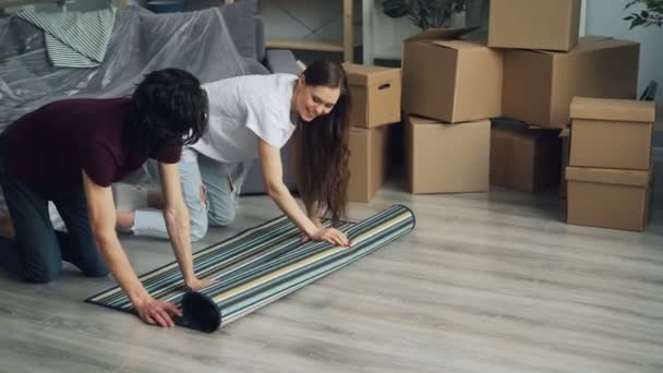 Hombre y mujer desplegando la alfombra en el suelo después de mudarse a un nuevo apartamento juntos — Vídeos de Stock