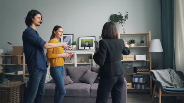 Wesoła para rozmawia z agentem nieruchomości patrząc na nowe mieszkanie w pomieszczeniu — Wideo stockowe