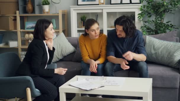 Junges Paar im Gespräch mit Makler beim Blick auf Dokumente und Wohnungsgrundriss am Tisch — Stockvideo