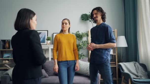 Мужчина разговаривает с риэлтором, пока жена смотрит на новую квартиру, стоящую в помещении вместе — стоковое видео