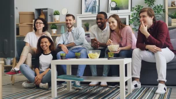 Jovens homens e mulheres assistindo TV rindo comendo pipocas no apartamento juntos — Vídeo de Stock