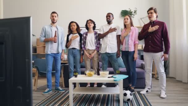 Fãs de futebol cantando hino com a mão no peito em pé em casa assistindo TV — Vídeo de Stock