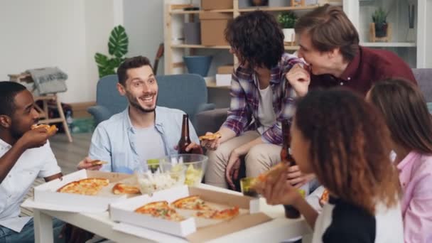 Молодые люди едят пиццу в чате и смеются над смешной вечеринкой в квартире — стоковое видео