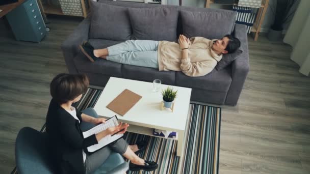 High angle view of teenage boy talking to psychologist lying on couch in office — Stock Video