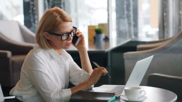 Зріла бізнес-леді робить мобільний телефонний дзвінок і використовує ноутбук в кафе — стокове відео