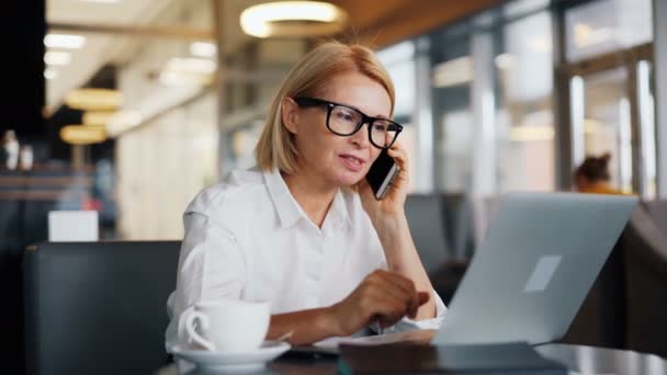 Alegre empresaria hablando en el teléfono móvil y utilizando el ordenador portátil en la cafetería — Vídeos de Stock