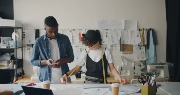 Diseñador de moda afroamericano usando teléfono inteligente cuando la niña mide la tela — Vídeos de Stock