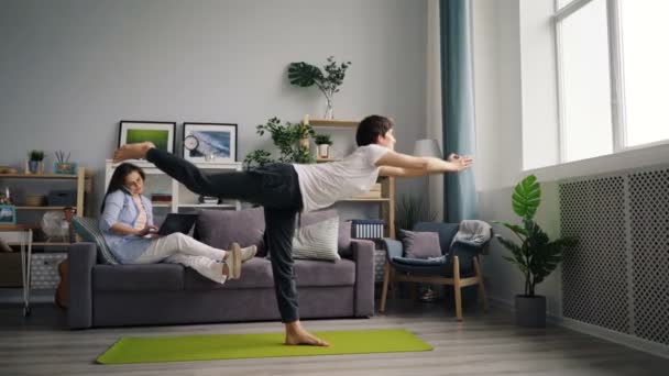 Asiatisk kille gör yoga övningar när fru pratar på mobiltelefon med hjälp av laptop — Stockvideo