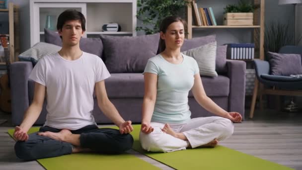 Genç bayan ve kocası evde meditasyon yapıyorlar. Lotus pozisyonunda minderlerin üzerinde oturuyorlar. — Stok video