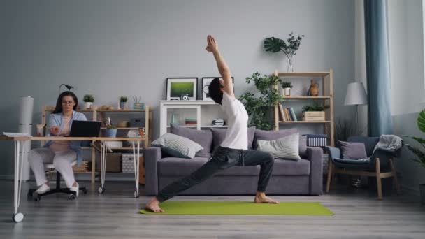 Man gör yoga asanas utbildning medan flickan arbetar med laptop på bordet hemma — Stockvideo