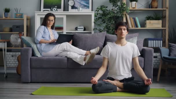 Asiatisk kille fokuserade på meditation medan hustru arbetar med dator hemma — Stockvideo