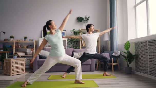 Aantrekkelijke jonge mensen meisje en man doen yoga thuis praktiserende asana's — Stockvideo