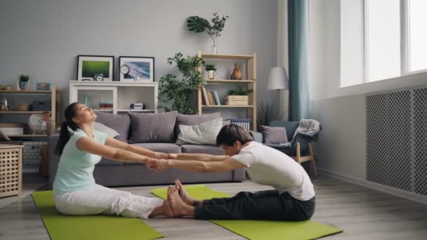 Чоловік і жінка роблять розтяжки, тримаючи руки, сидячи на килимках вдома — стокове відео
