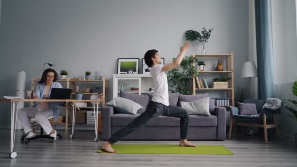 La moglie sta lavorando con il computer portatile a tavola mentre il marito sportivo esercita facendo yoga — Video Stock