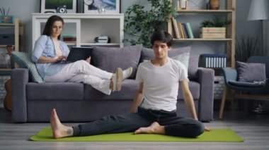 Esnek genç adam kanepede oturan dizüstü bilgisayar kullanırken kız arkadaşı yoga yapıyor