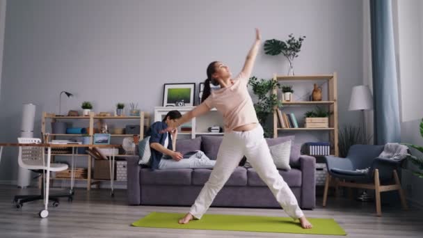 Chica haciendo asanas de yoga mientras el hombre trabaja con el ordenador portátil hablando por teléfono móvil — Vídeos de Stock