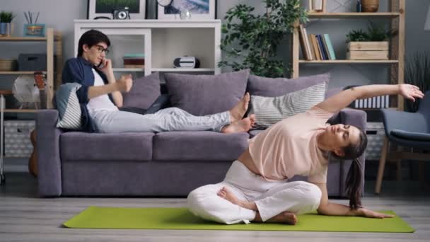 Молода жінка практикує йогу вдома, поки хлопець фрілансер розмовляє по телефону — стокове відео