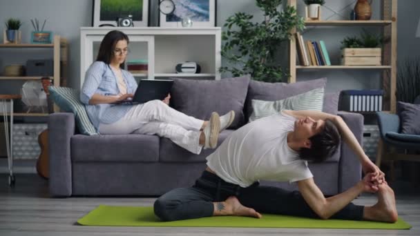 Азіатський молодий чоловік практикуючих йогу будинку в той час як його дружина за допомогою ноутбука на дивані — стокове відео