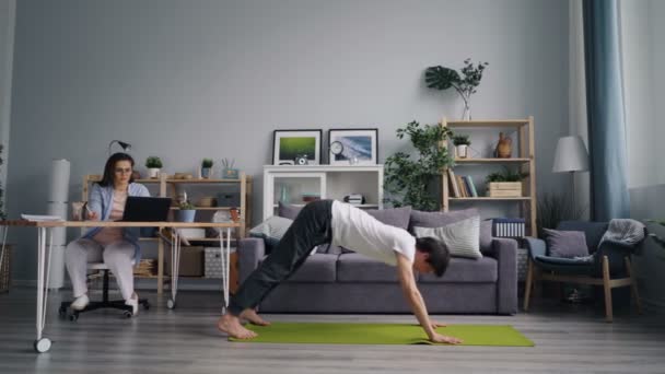Estudiante femenina trabajando con laptop estudiando cuando su marido hace yoga en casa — Vídeos de Stock