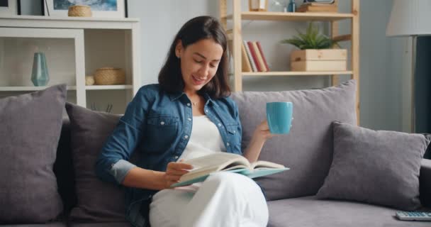 Movimento lento de menina atraente leitura livro segurando xícara de café em casa — Vídeo de Stock