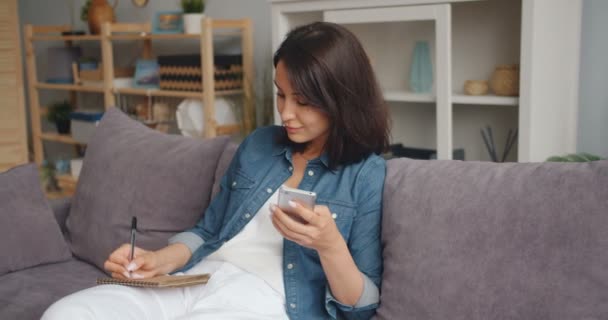 Slimme vrouw schrijven in notebook met behulp van smartphone glimlachend zittend op sofa thuis — Stockvideo