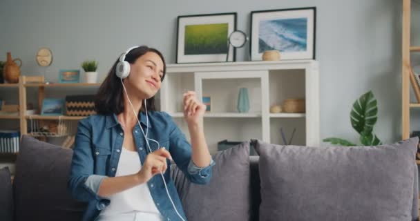 Lento movimento da menina em fones de ouvido dançando ouvir música sentada no sofá — Vídeo de Stock