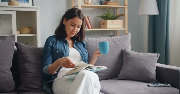 Jovencita inteligente leyendo libro sosteniendo la taza de té sentada en el sofá en el apartamento — Vídeos de Stock