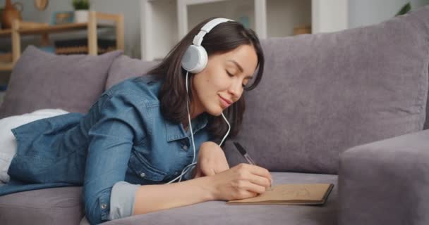 Mooie vrouw in een koptelefoon luisteren naar muziek en tekening foto in notebook — Stockvideo